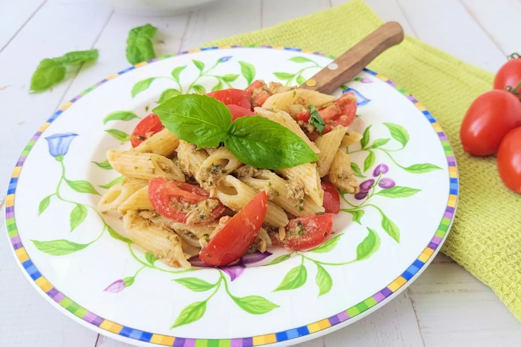 pasta fredda pesto tonno e pomodorini, piatto di pasta fredda con sullo sfondo uno strofinaccio verde, dei pomodorini e delle foglia di basilico